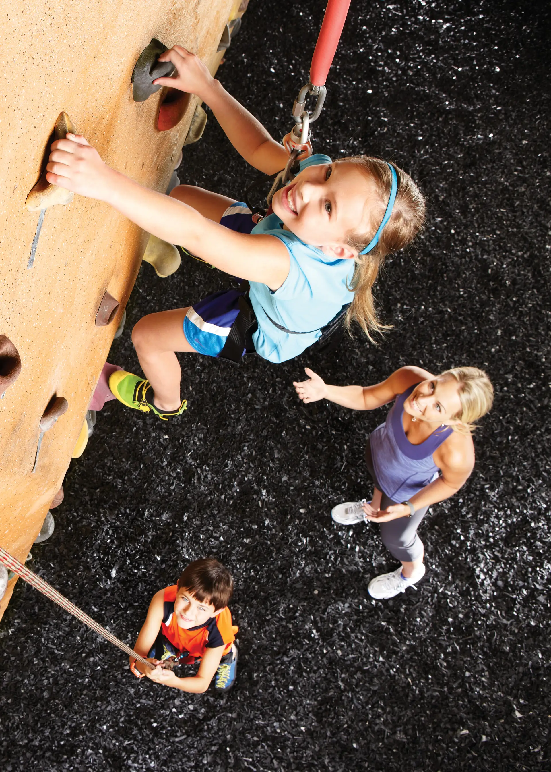 A smiling child in a harness climbing on a rock wall while two other smiling people watch.