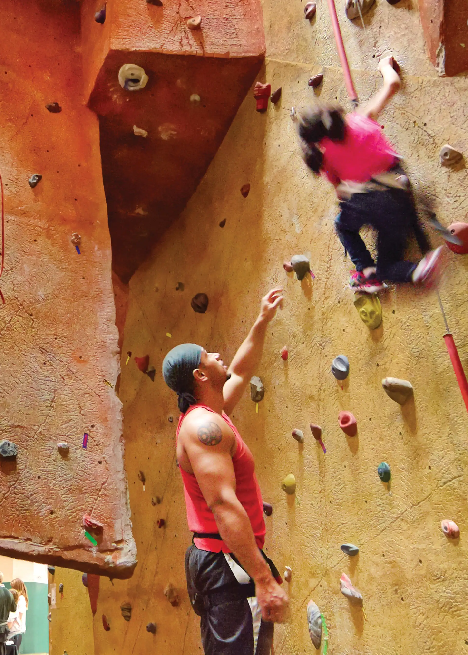 Person in harness climb up an indoor rock climbing wall while an instructor looks on.