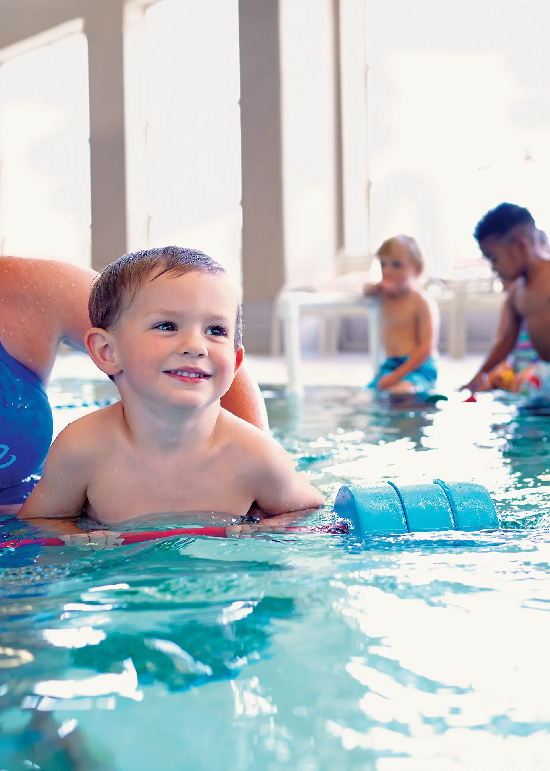 A smiling child swimming in an indoor pool assisted by a flotation device and an adult.