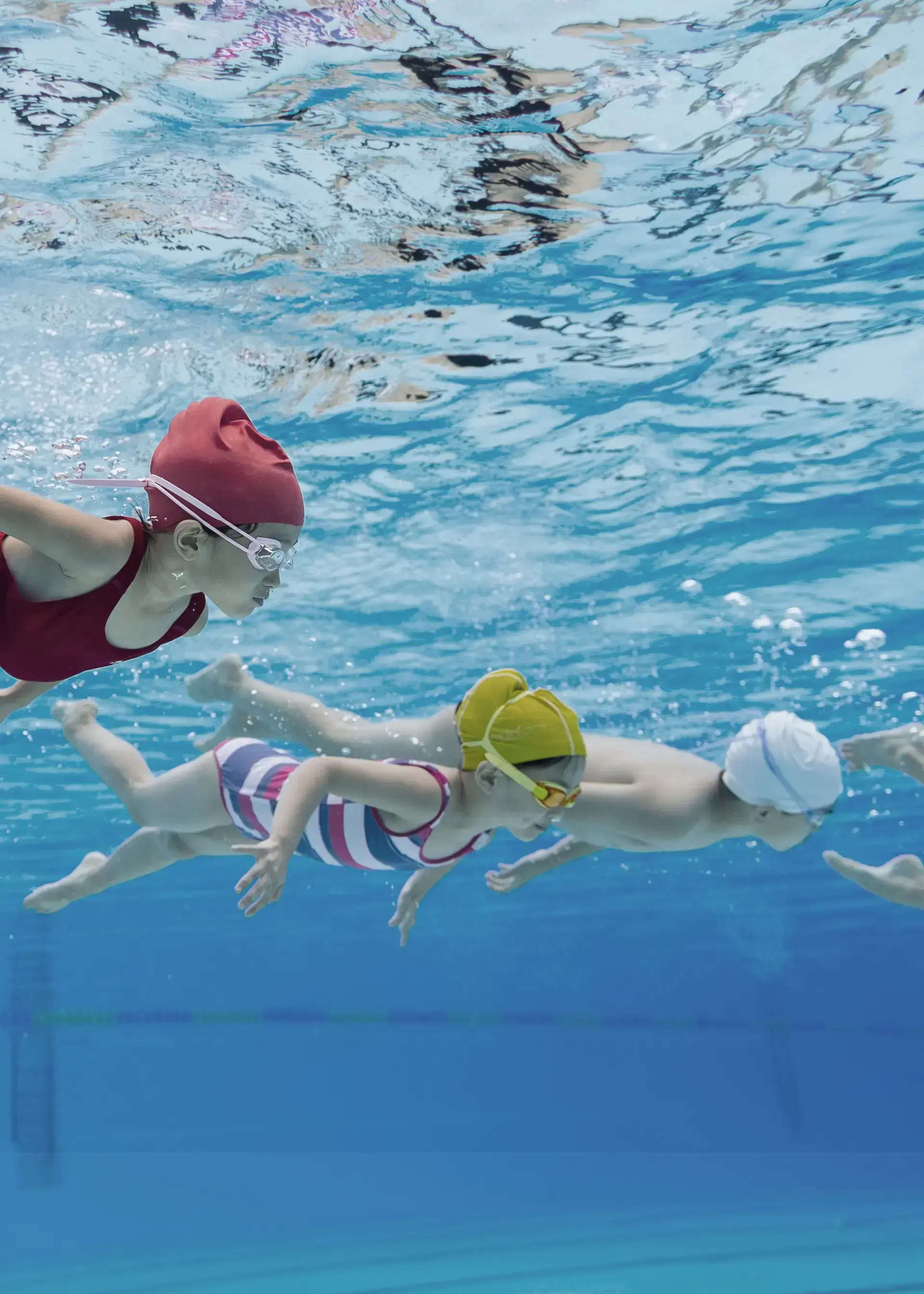 Three kids swimming under water in swimsuits, goggles and swim caps.