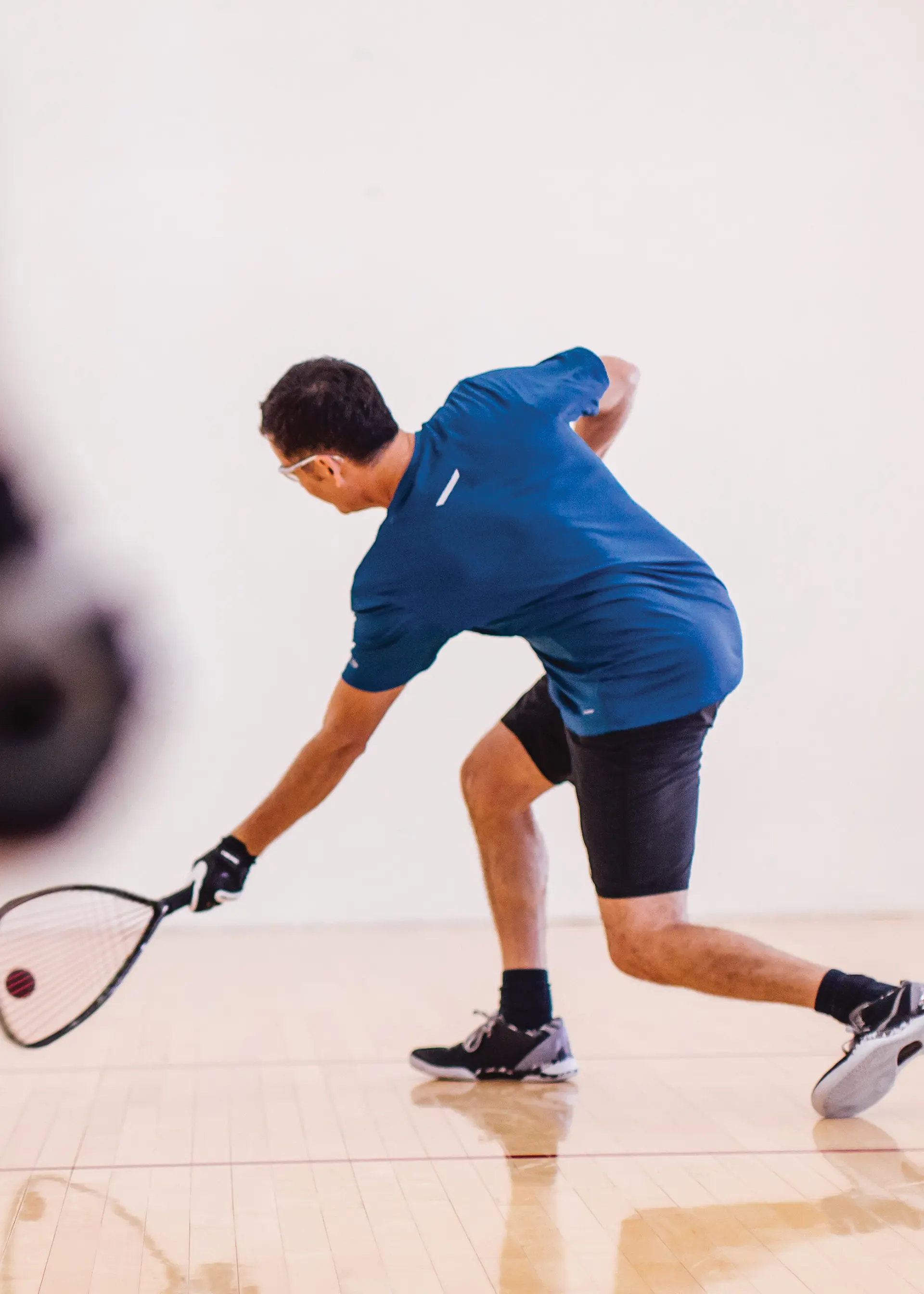 A person playing squash and reaching for a ball on an indoor court.