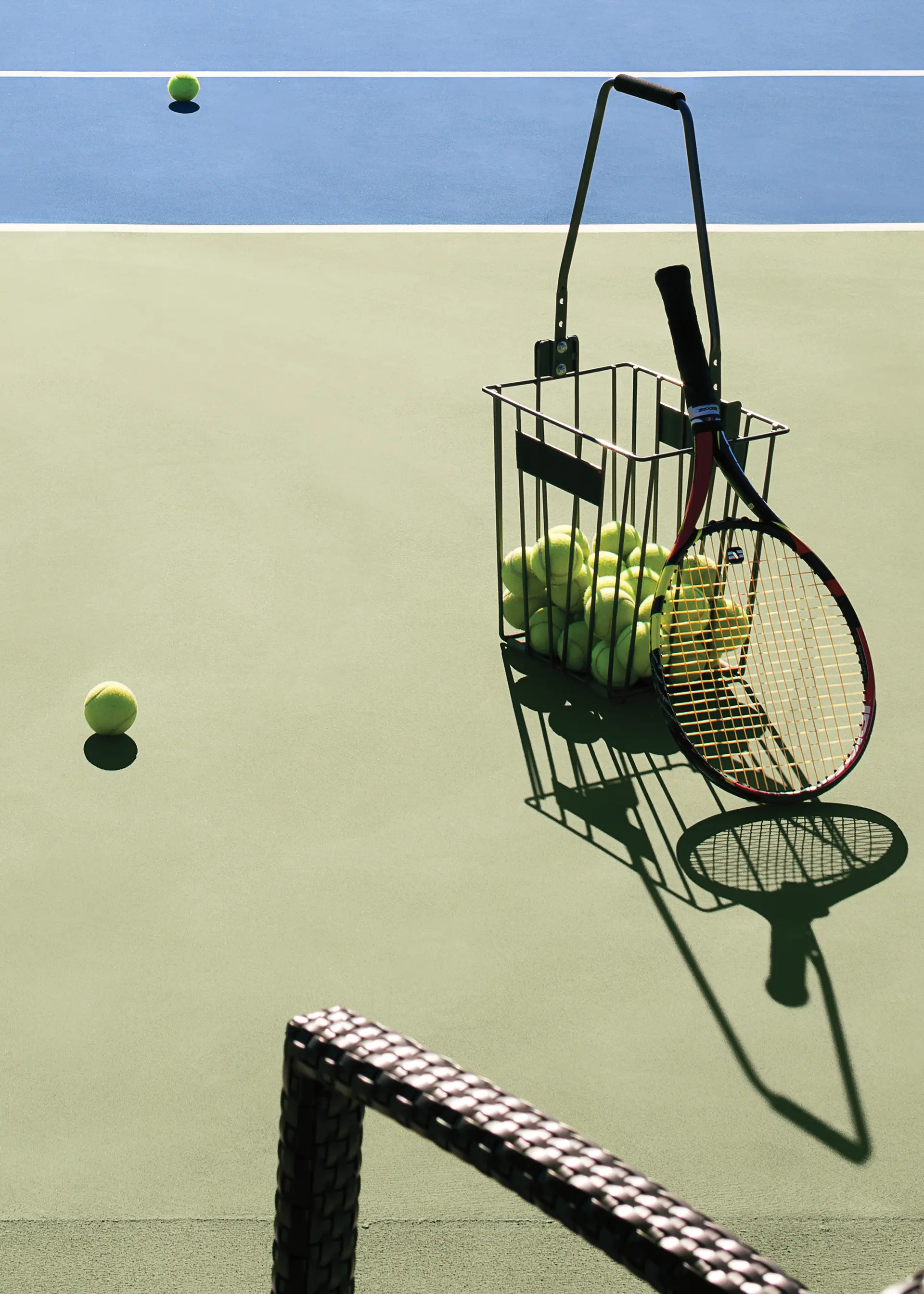 A tennis racket and a basket of tennis balls on a court.