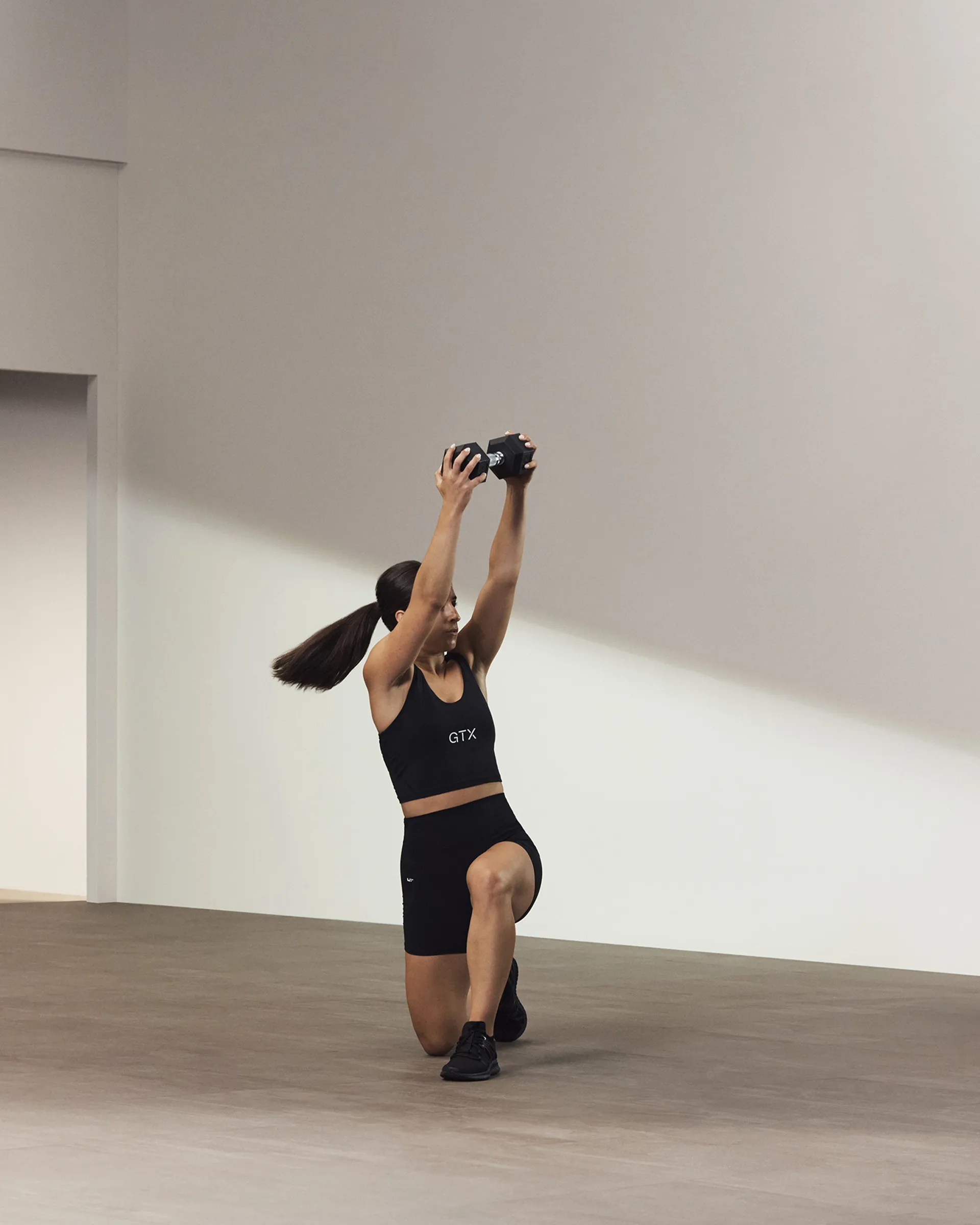 A Life Time member on one knee and holding a dumbbell overhead.