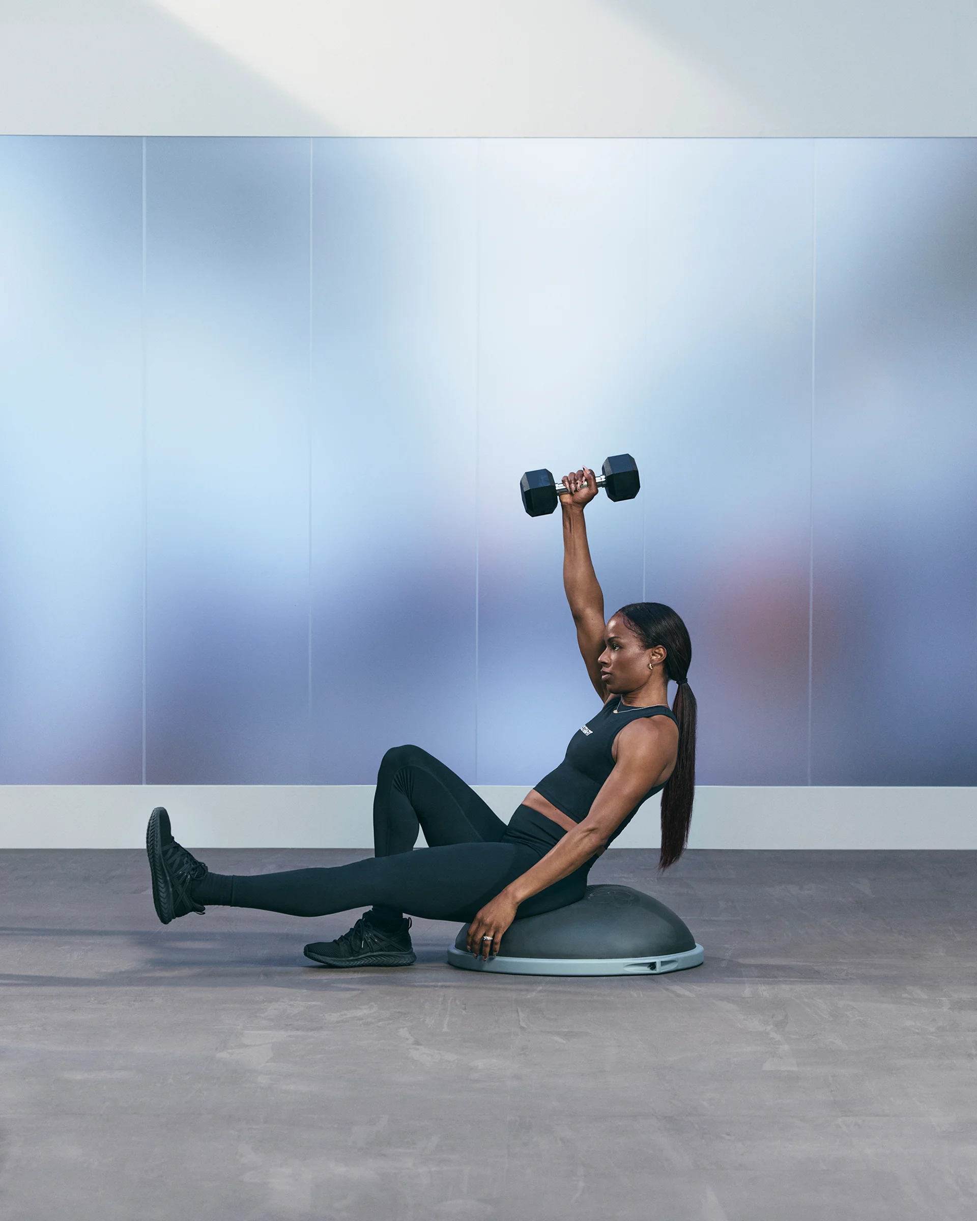 A Life Time member sitting on a BOSU with one leg stretched out and one arm up holding a dumbbell.