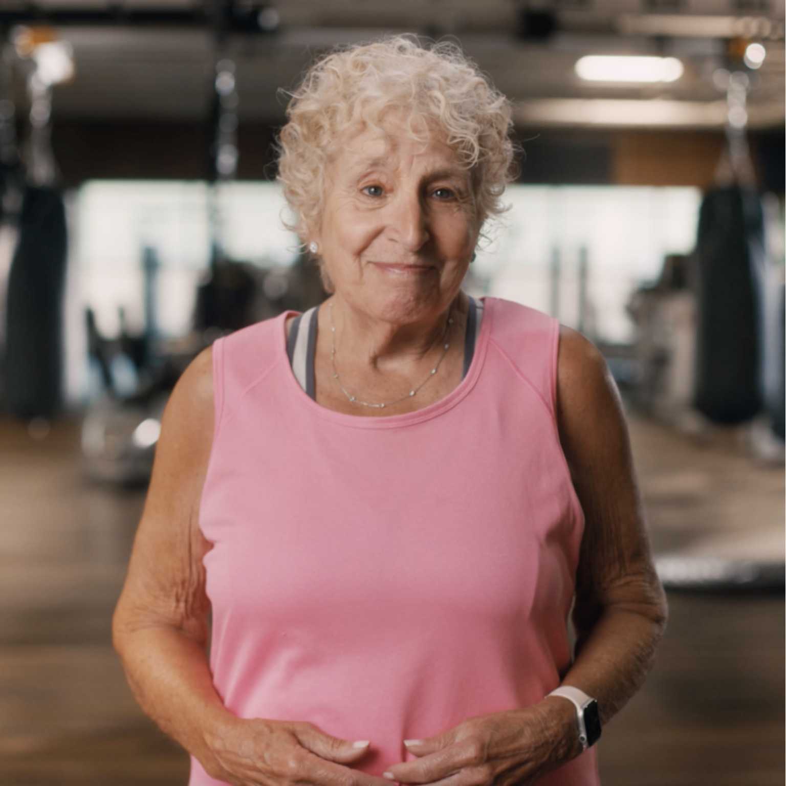 A woman in a pink tank top smiles at the camera with exercise equipment behind her. 