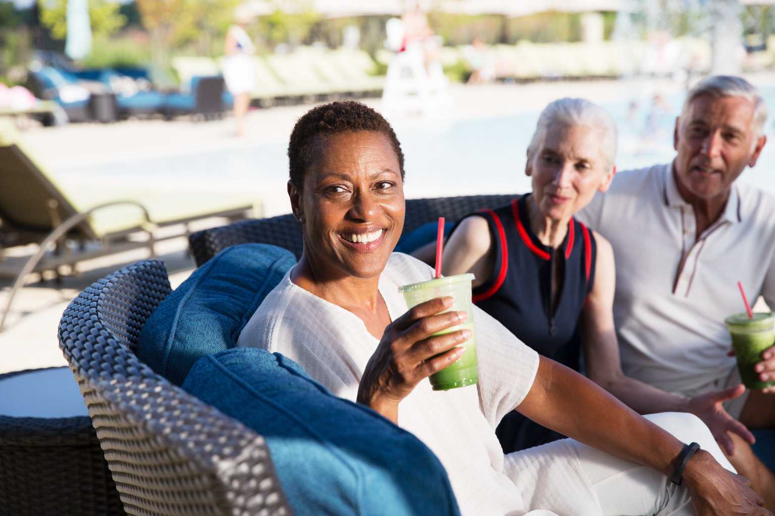 Two women and a man sitting outside by the pool holding smoothies. 