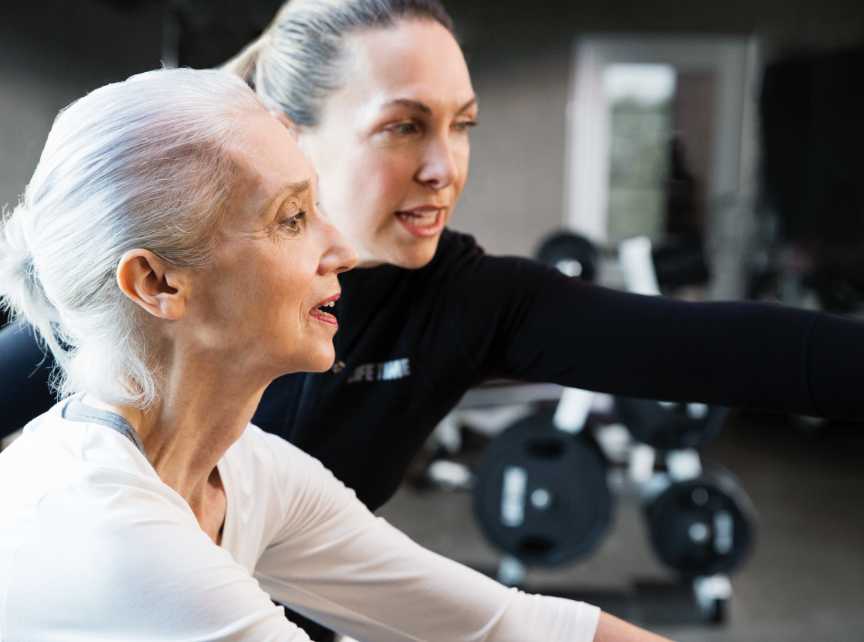 A Life Time personal trainer helps an woman on a piece of exercise equipment.