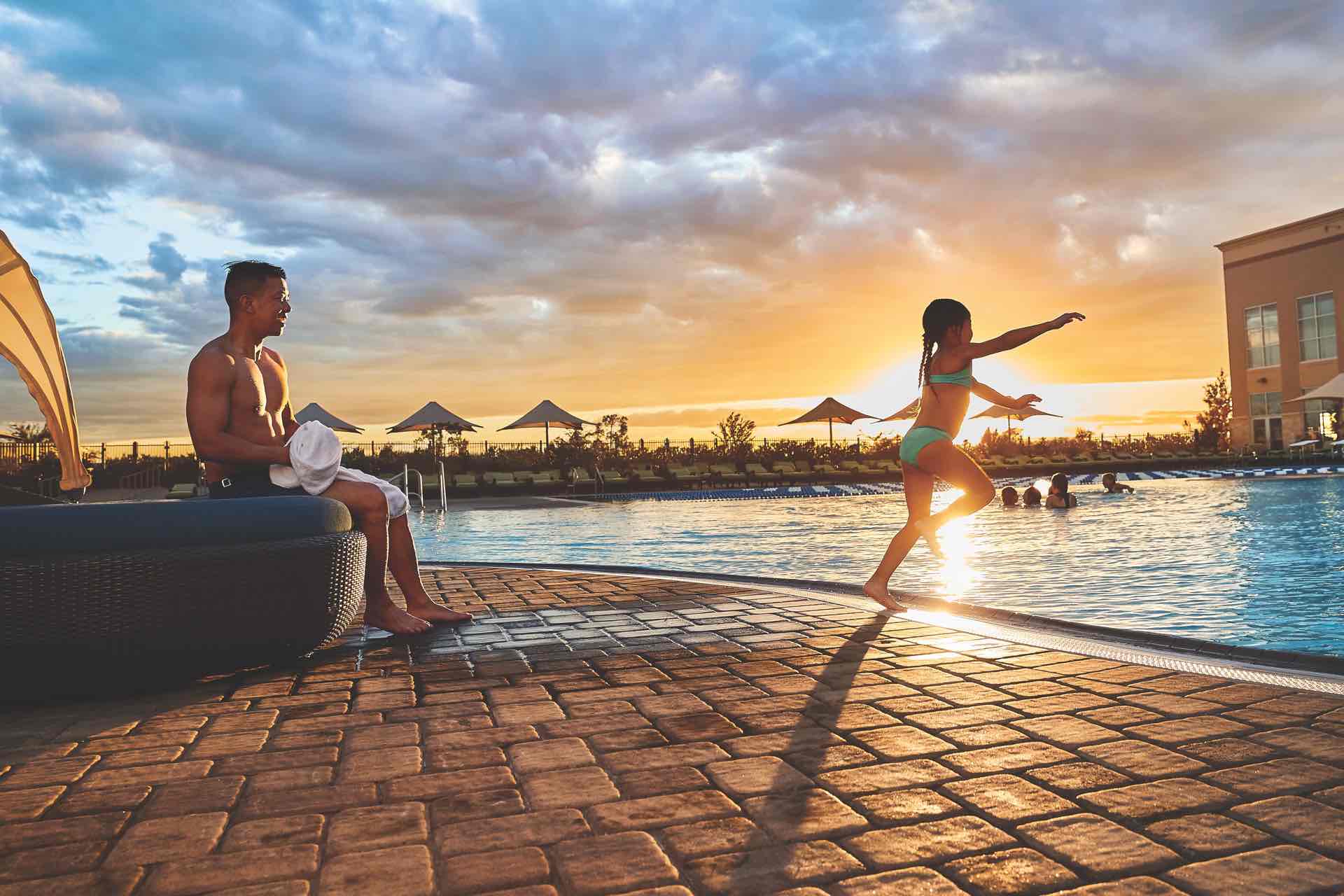 Man watching a female child jump into an outdoor pool as the sun sets at Life Time