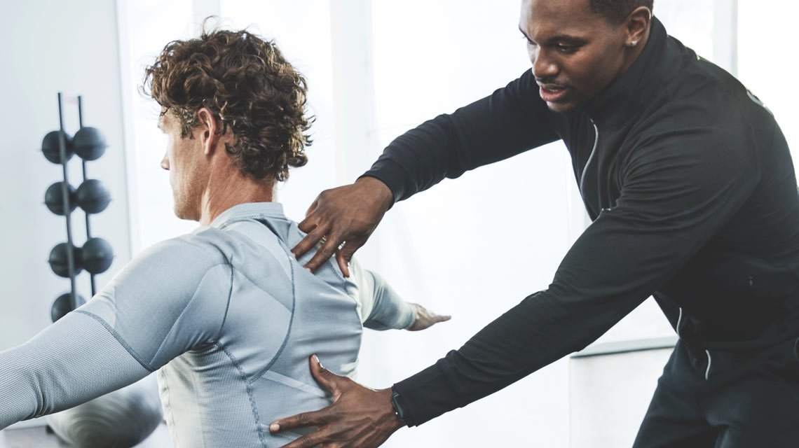 Life Time Health and Wellness trainer places his hands on the back of a client who is sitting with his arms out