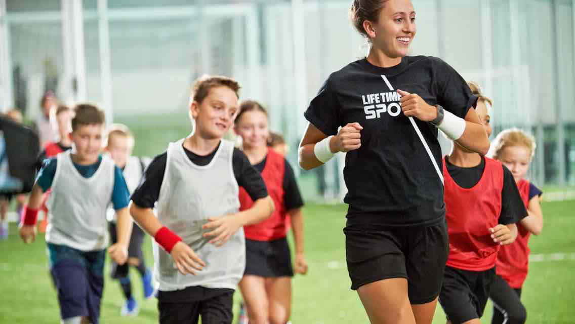 A soccer coach jogging on a turf field followed by six children in red and white soccer practice jerseys