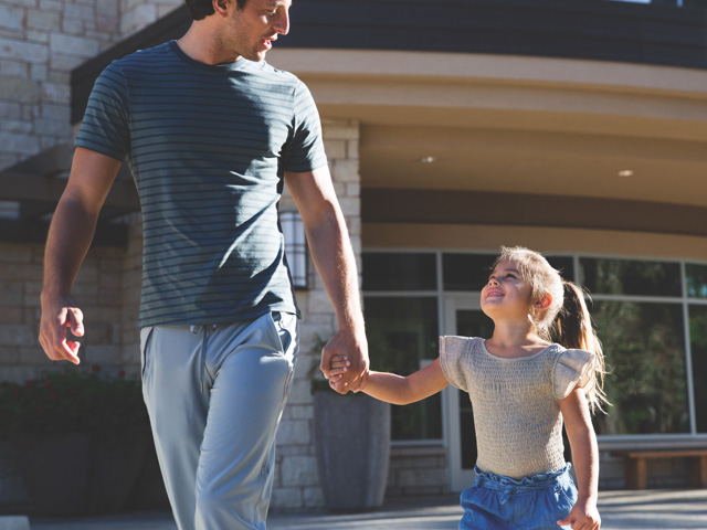 A young girl holds her dads hand and smiles up at him while exiting a Life Time location
