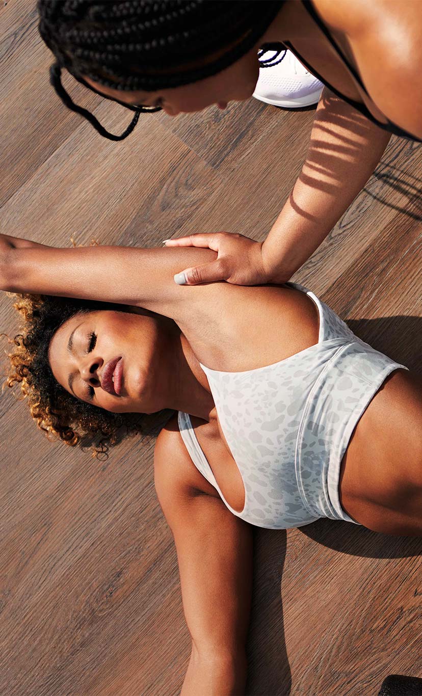 a woman lays on the ground and has a trainer assist in a stretch
