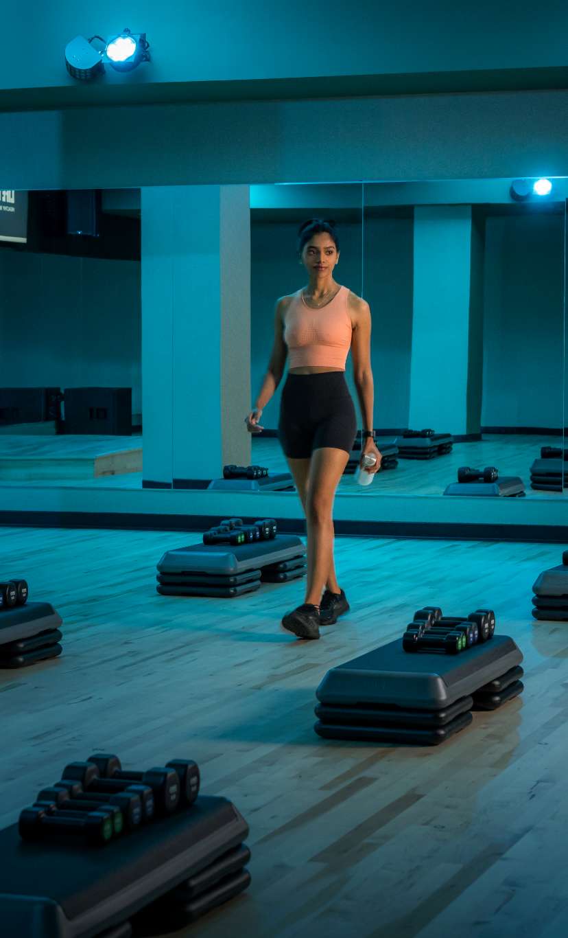 a woman walks through rows of aerobic steps with barbells in a blue lit group fitness space