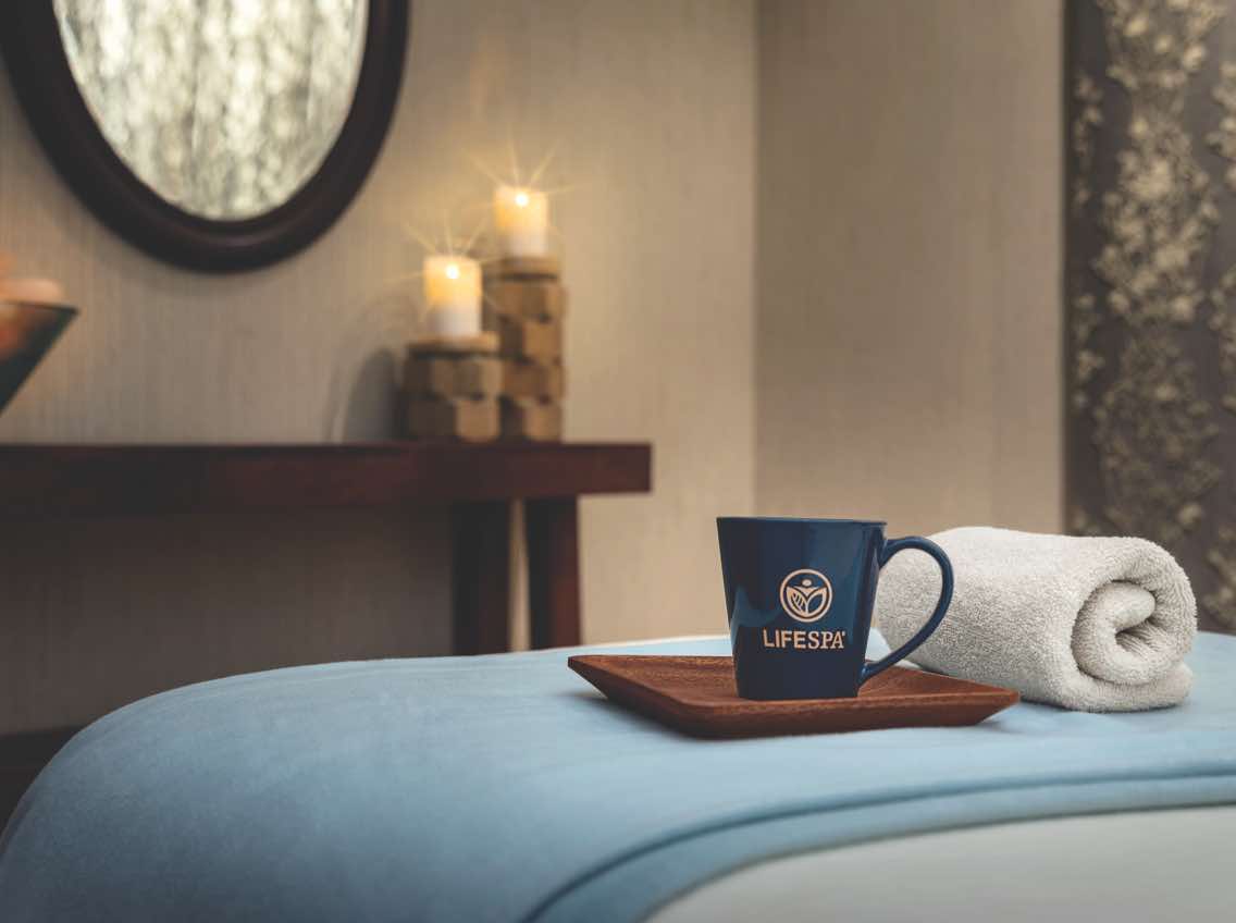 A rolled white towel and a mug with a LifeSpa logo sit atop a blanket-covered massage table