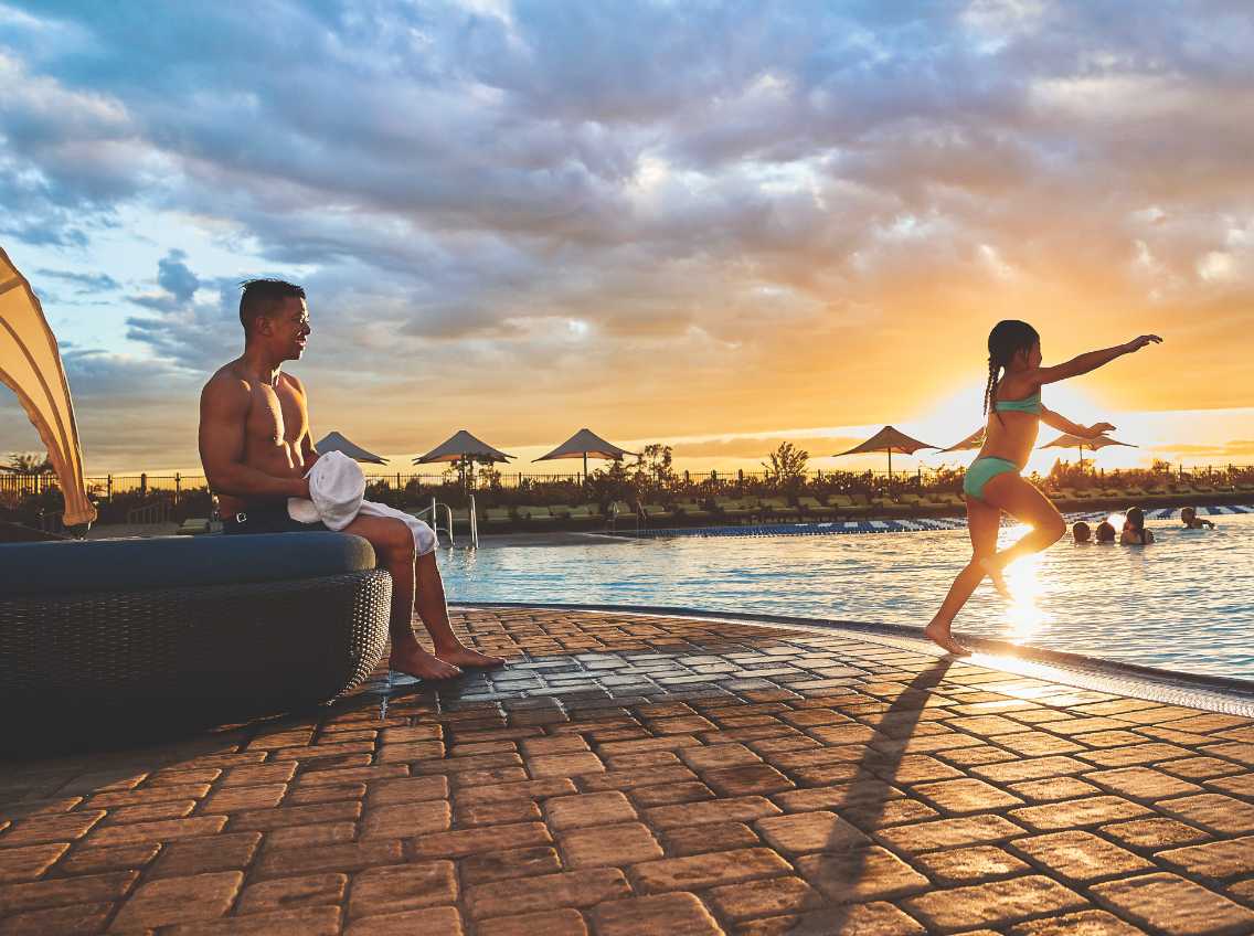 Girl jumping into a pool at sunset with her dad in a lounge chair behind her. 