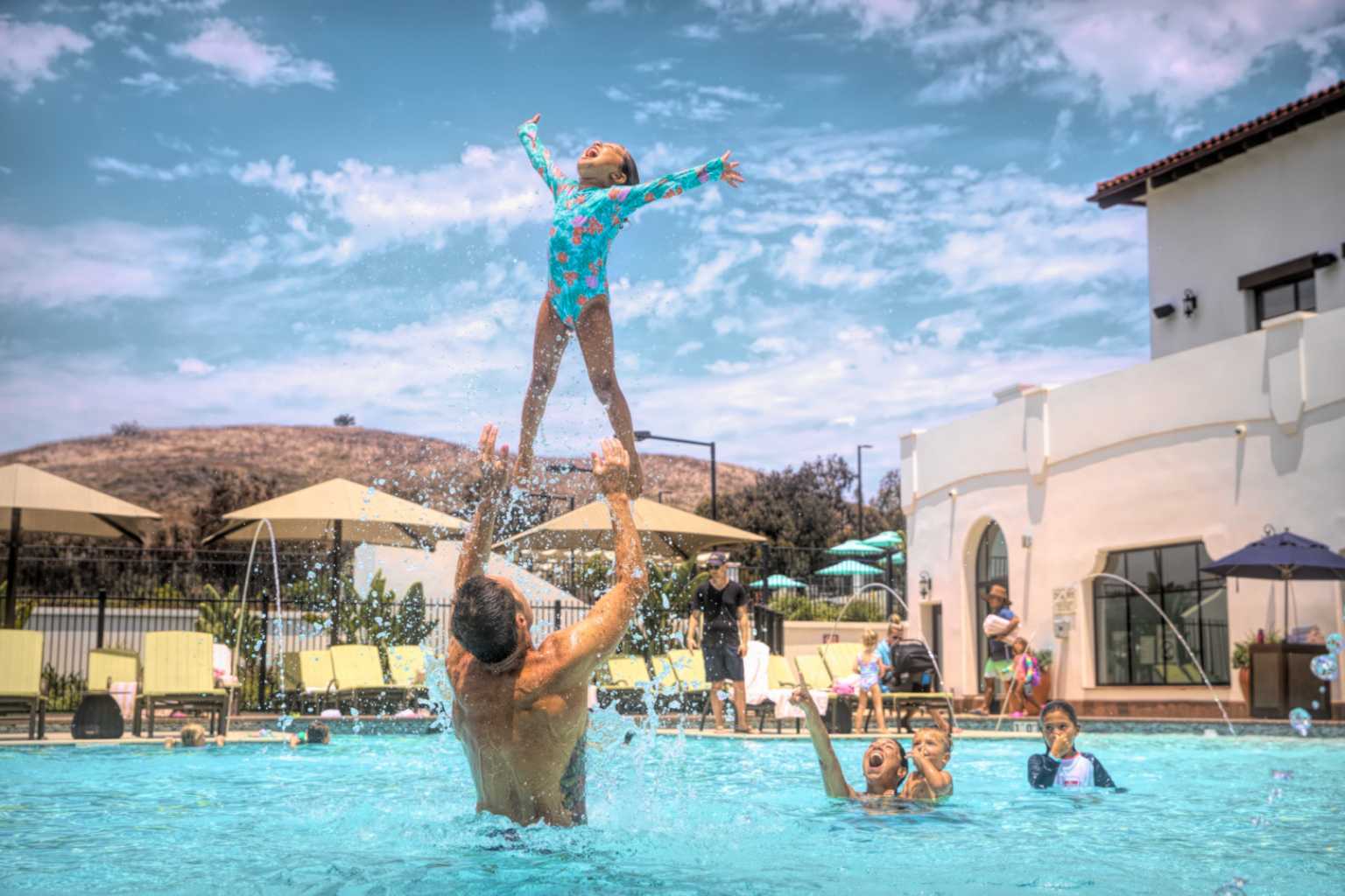 a dad throws his daughter into the air, while playing in an outdoor pool with other children