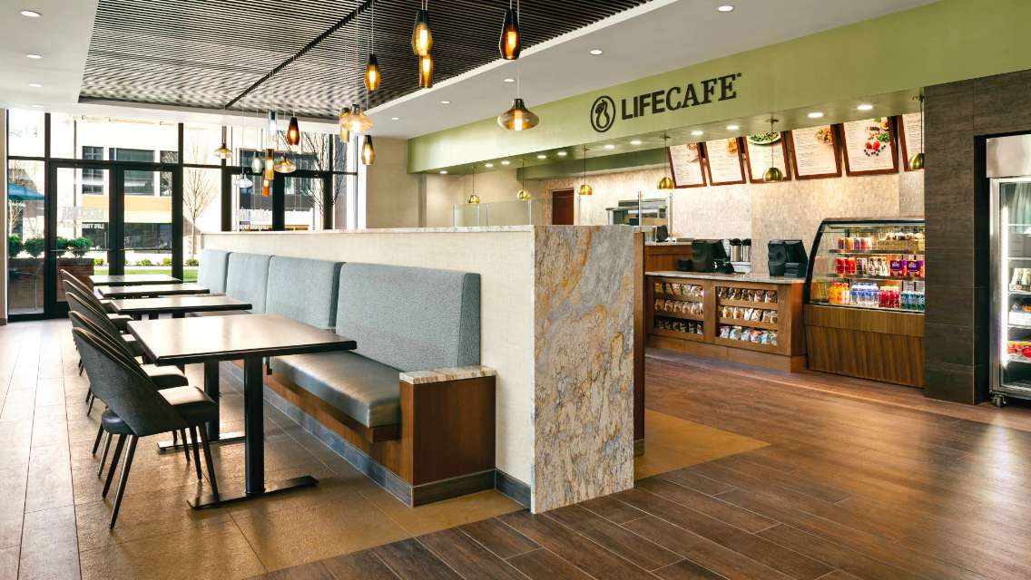 An architectural shot of LifeCafe with the walk-up counter, menu boards, retail food displays and tables and chairs for seating. 