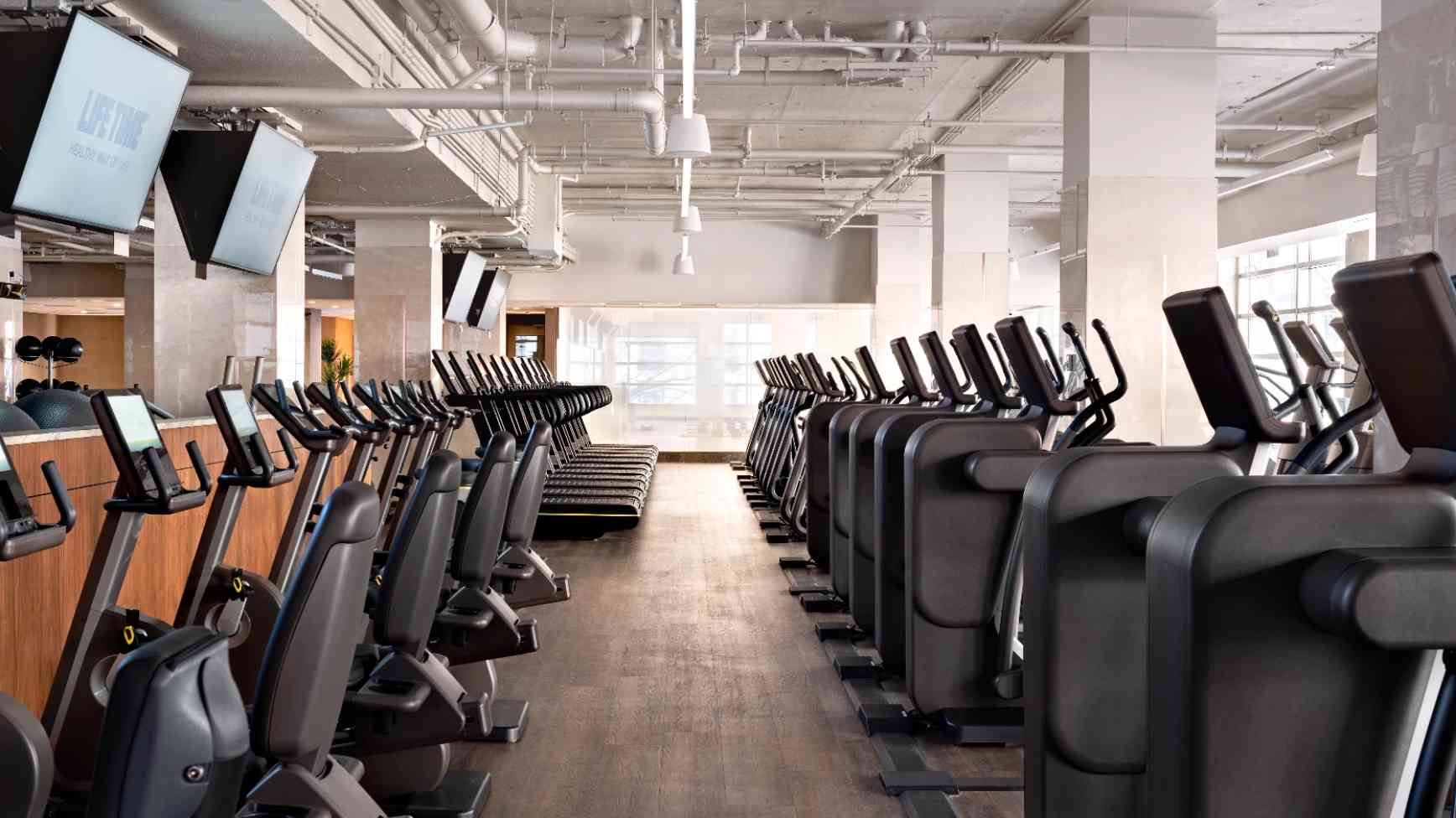 Workout floor space with high ceilings, wood floor, large flat-screen TVs. Long rows of upright bikes, treadmills and ellipticals within natural and artificial lighting.