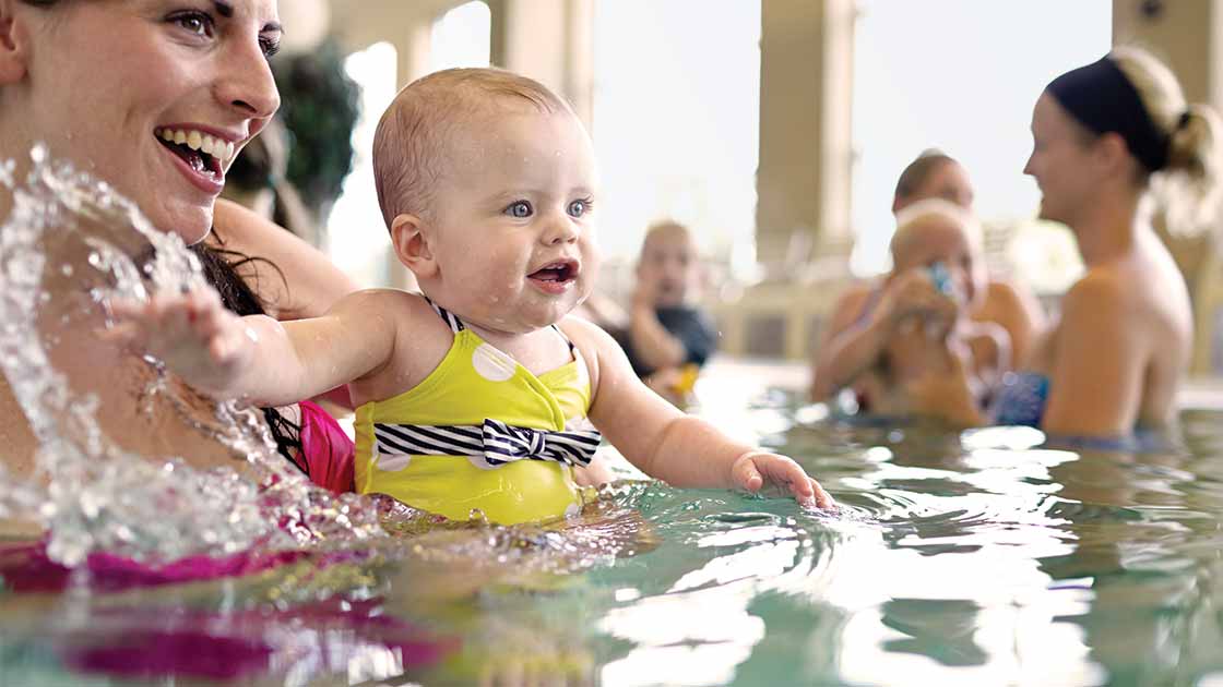 A baby girl splashing water by a woman in a baby swim class