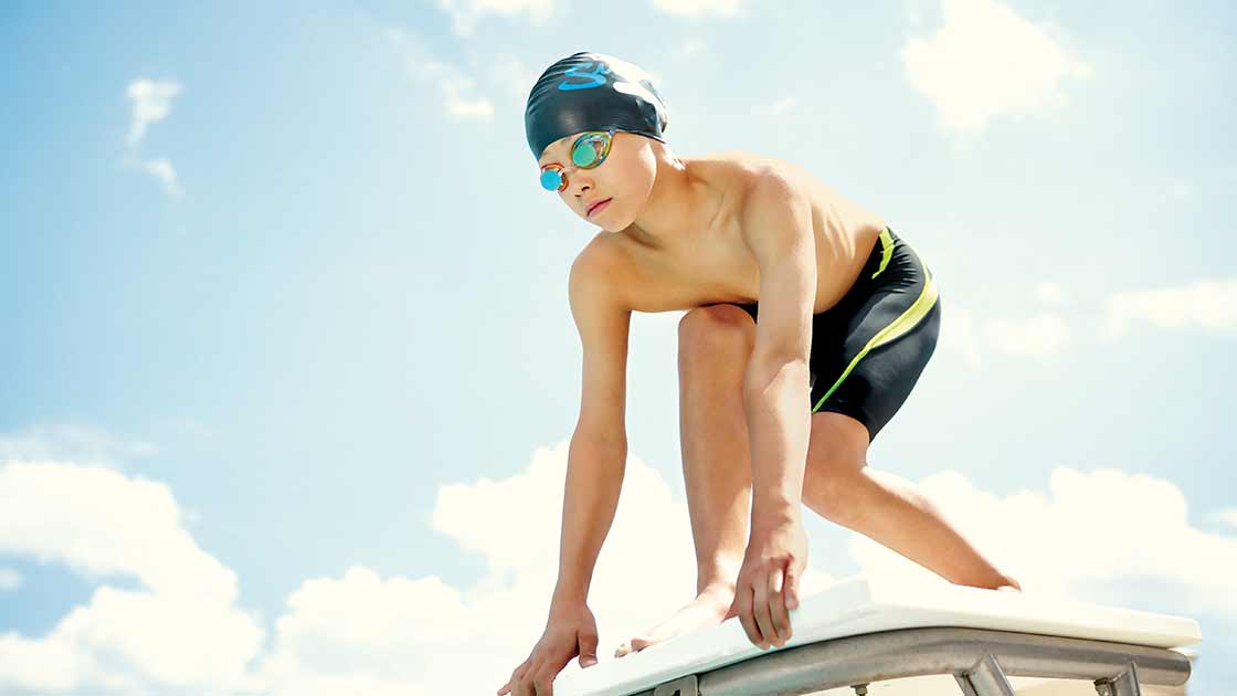 A boy wearing a swim cap and goggles preparing to dive into an outdoor pool