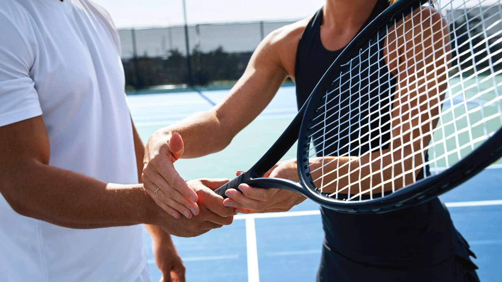 -	Person helping another person to properly hold a tennis racquet on an outdoor tennis court. 