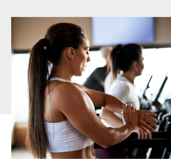 A Caucasian woman with long brown hair in a ponytail stands on a treadmill, checks her watch.