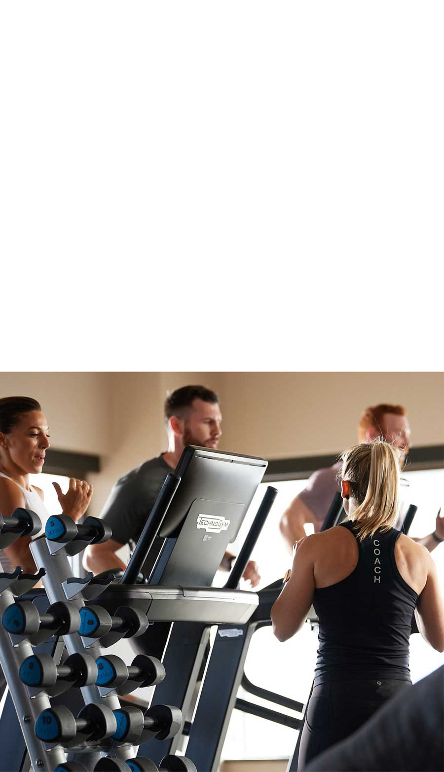 Three GTX members run on treadmills in a studio filled with beautiful morning light, a weight rack in the foreground. Coach Danica stands in front of them, her back to the camera, offering encouragement. 