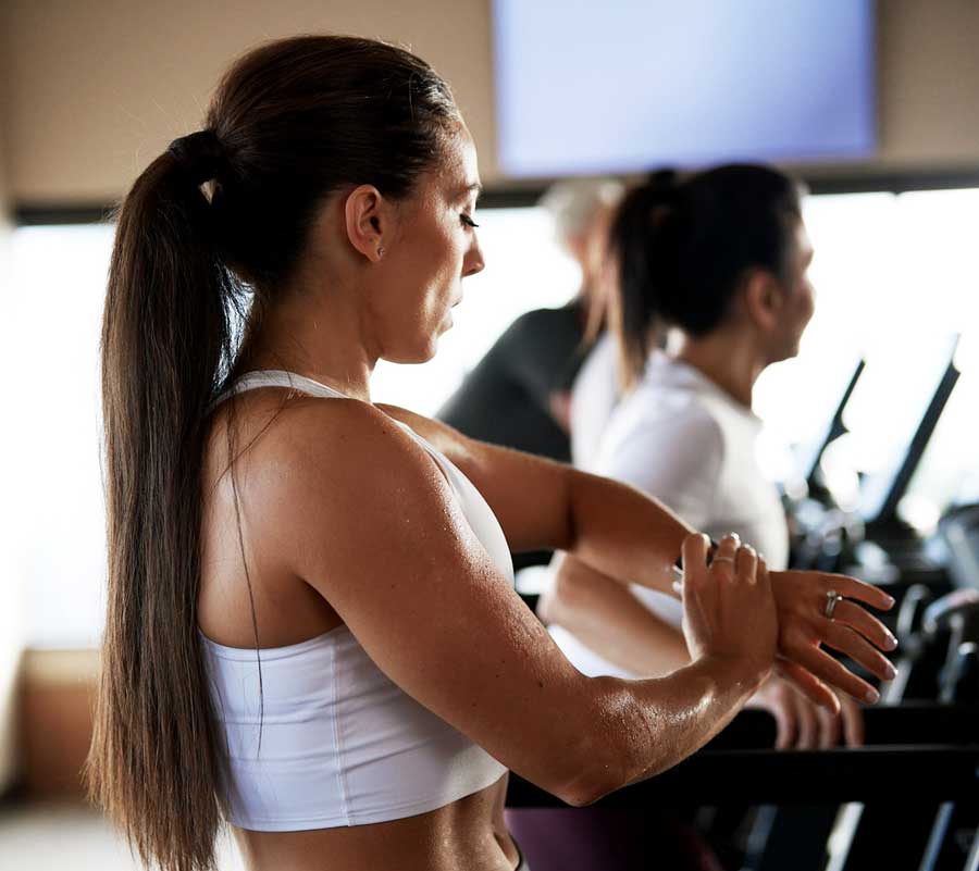 A Caucasian woman with long brown hair in a ponytail stands on a treadmill, checking her watch.