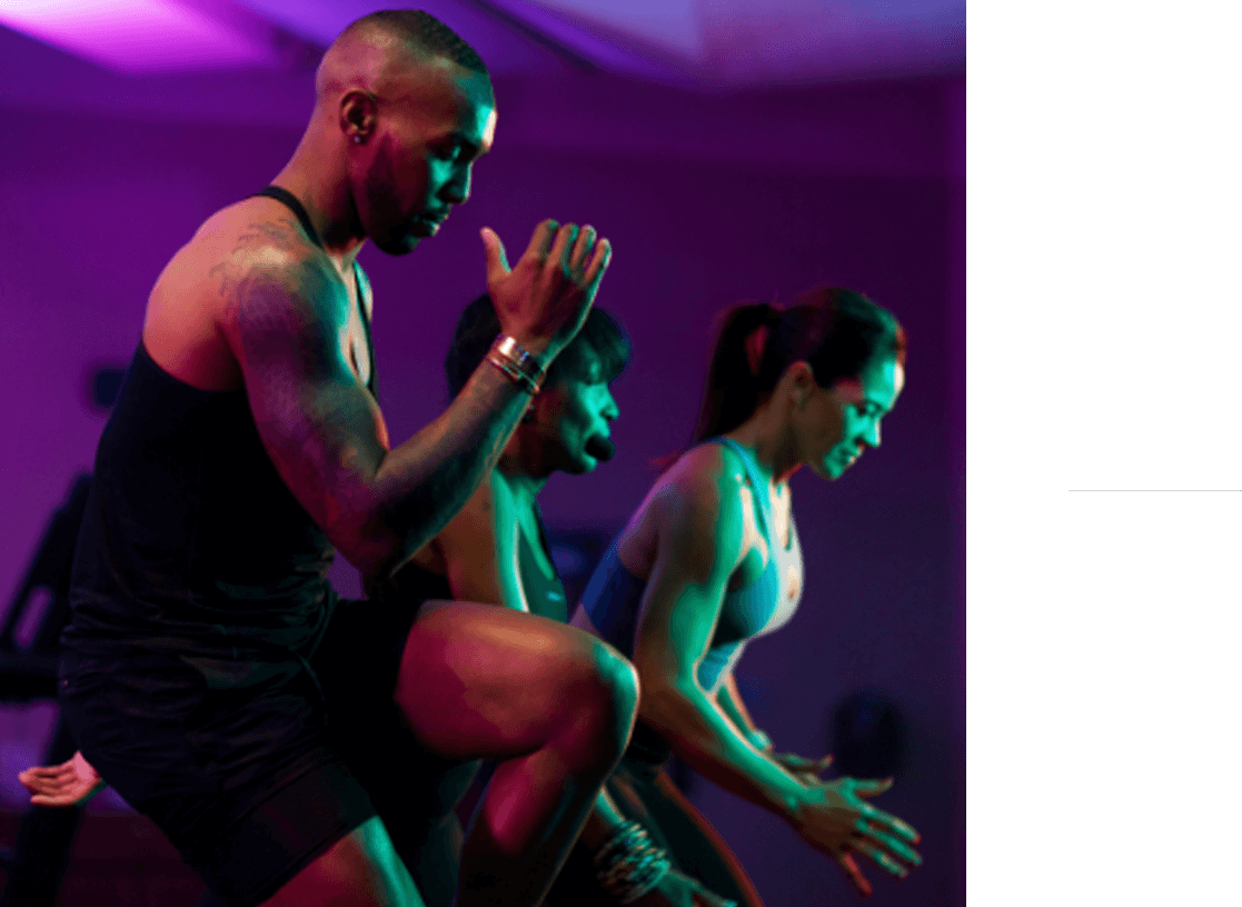 A side angle shot of a Black man from the knees up in an Ultra Fit class. He is exceptionally fit, with a full sleeve tattoo on his right arm, and is touching his right elbow to his left knee.