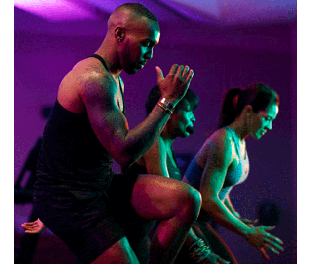 A side angle shot of a Black man from the knees up in an Ultra Fit class. He is exceptionally fit, with a full sleeve tattoo on his right arm, and is touching his right elbow to his left knee.