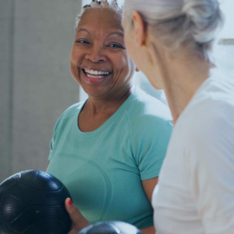 Two older women share a smile while working out with medicine balls