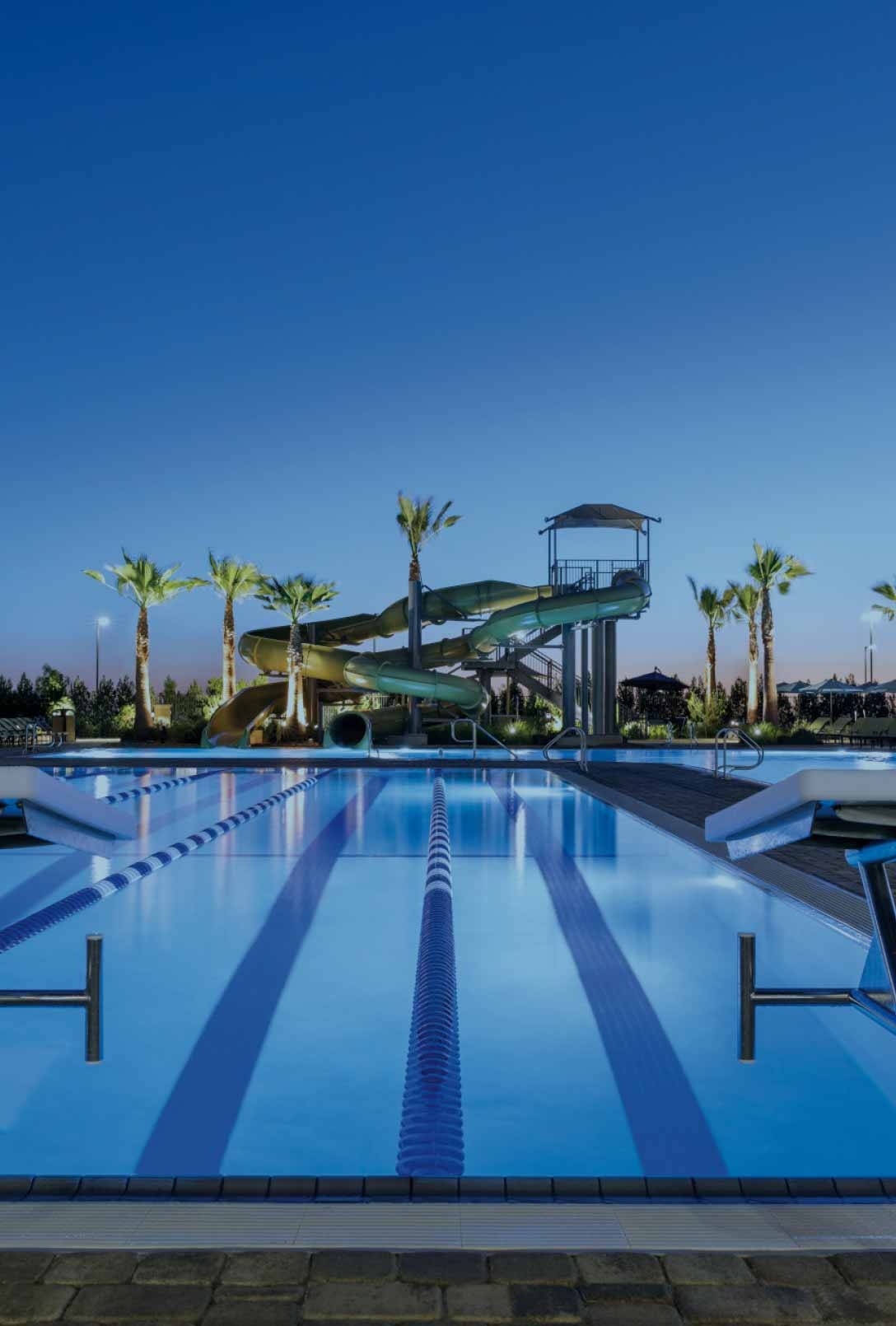 An outdoor lap pool featuring starting blocks at dusk, with waterslides in the background