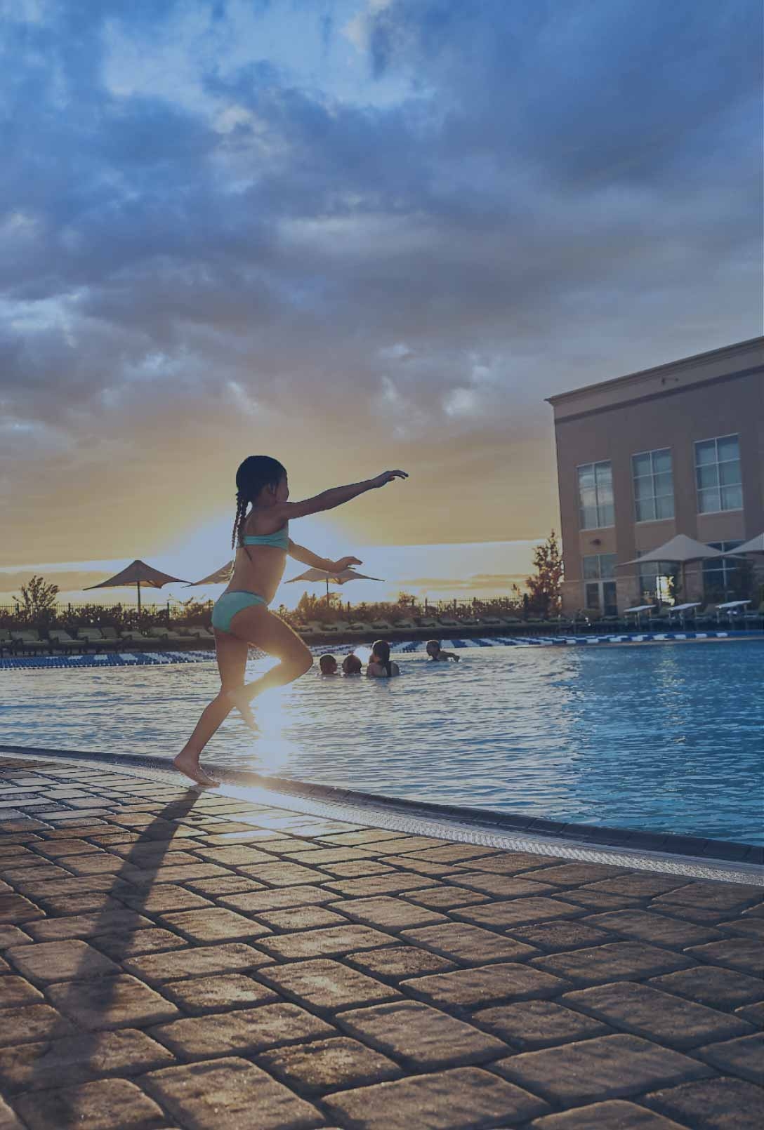 A dad watches his daughter jump into a life time outdoor pool at sunset