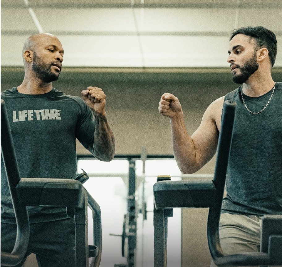 A trainer and his client share a fist bump in a celebratory moment. The two men stand side by side on treadmills as if they just finished working out together.