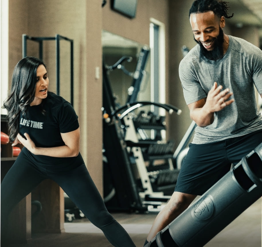 A female trainer models correct form for a sideways lunge for her client. He is smiling despite the additional challenge of using a Viper, a weighted cylinder that's used for strength training.