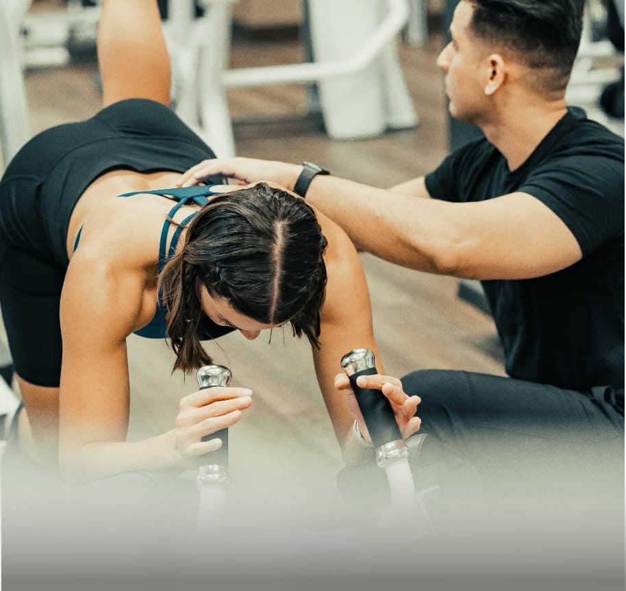 A male trainer working out with a client. She is prone on a piece of exercise equipment kicking her heel up to the ceiling. He lightly touches her back to help her activate the right muscles.
