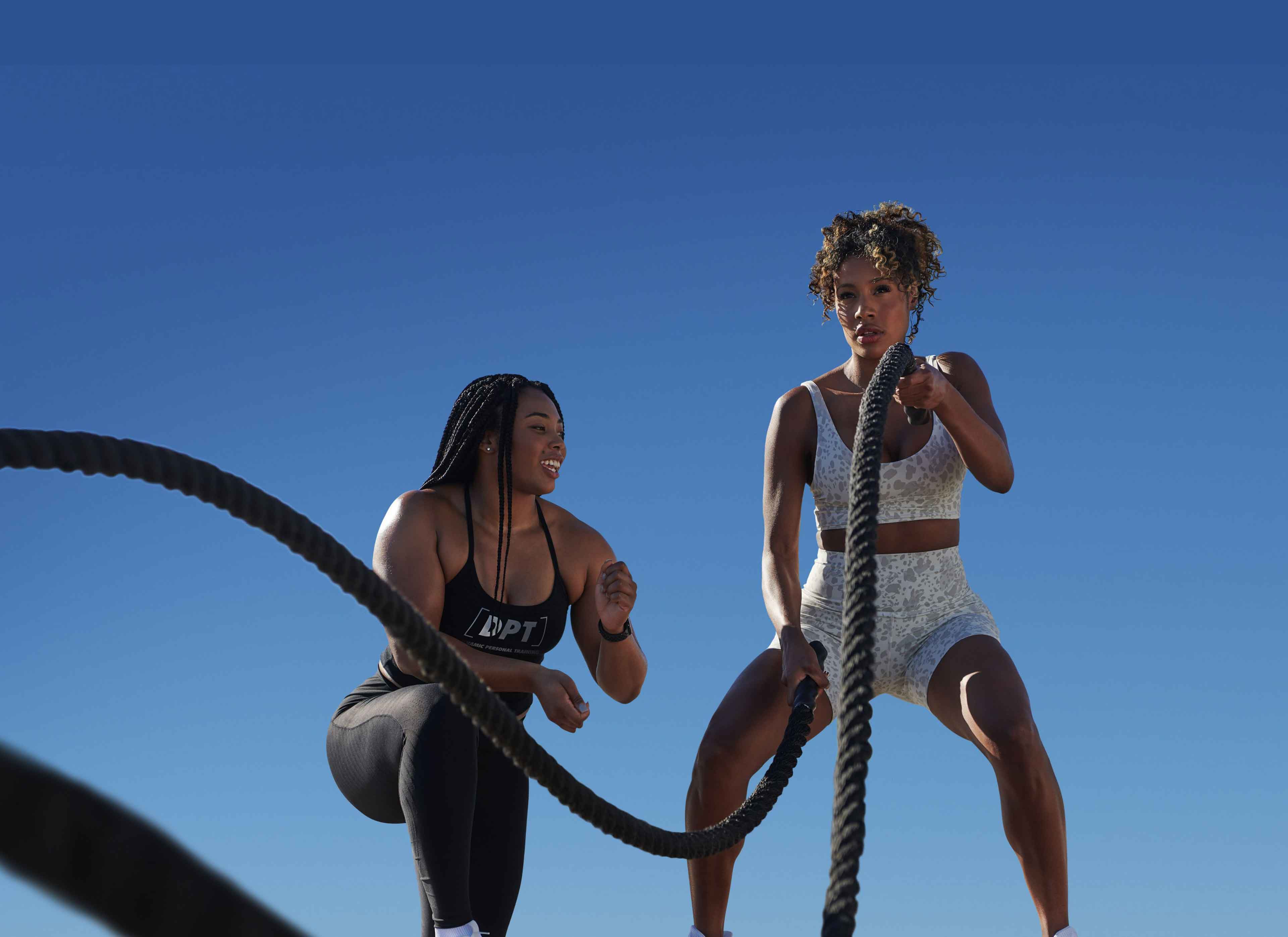 A female client works the battle ropes as her trainer kneels alongside her giving her encouragement.