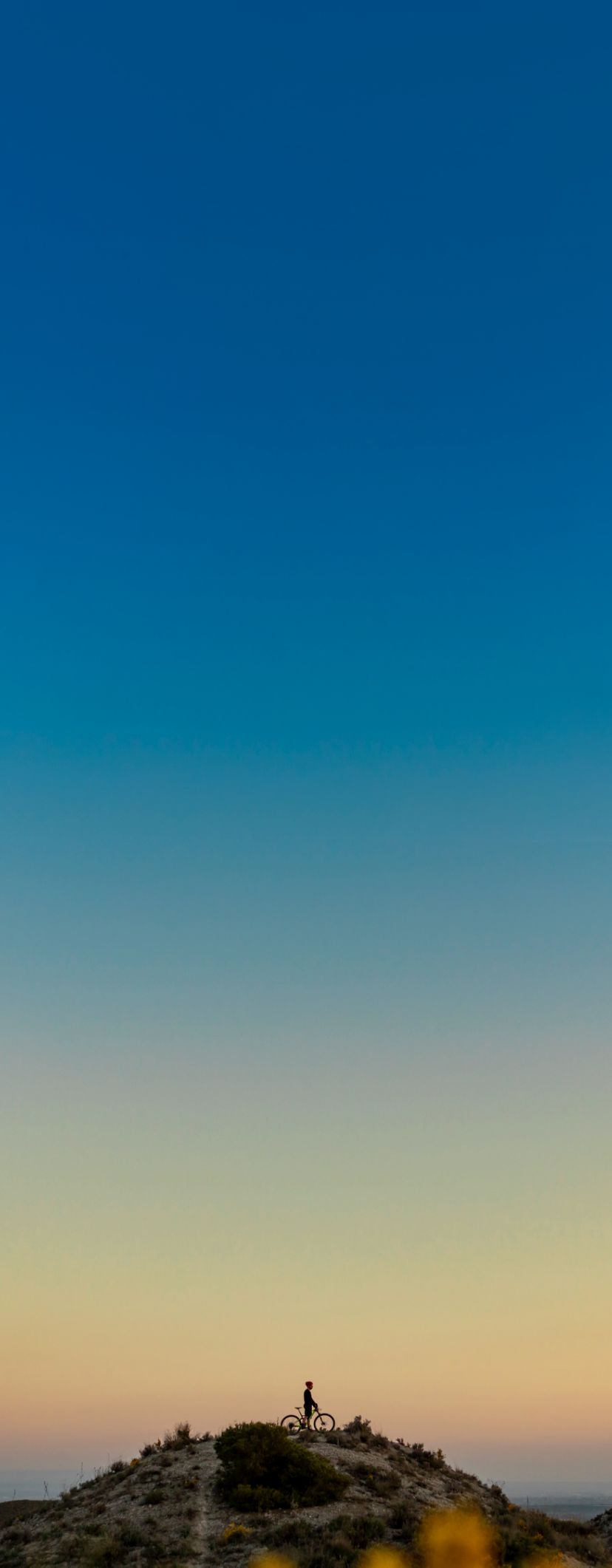 the silhouette of a man on a bike at the top of a mountain with a sunset behind him.